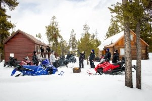 A far shot of Snowmobiles outside of a cabin with various people.