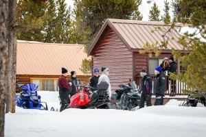 Snowmobiles outside of a cabin with various people.